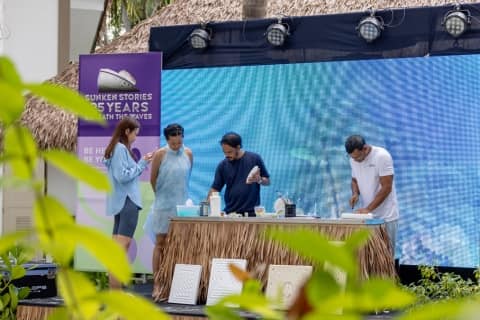 A group of people standing around a table, with an artist demonstrating the intricate craft of carving coral stone using discarded styrofoam.